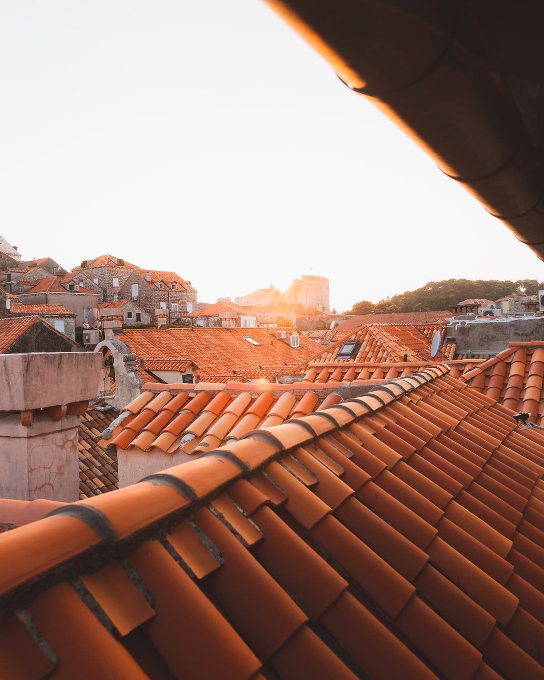 Tejados de tejas rojizas con chimeneas, ventanas y antenas, edificios de piedra al fondo y luz de sol junto al horizonte.